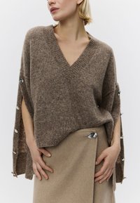 Pulover maro din amestec de lână, cu decolteu în V, croială lejeră și crăpături laterale, prevăzut cu nasturi de culoare argintie pe mâneci. Asortat cu o fustă bej.
