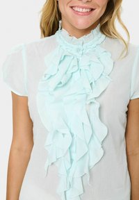 Blusa azzurra chiara con maniche corte, caratterizzata da un design frontale con volant e un alto colletto arricciato. Il tessuto appare leggero e con una texture.