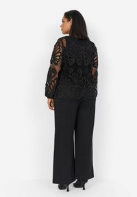 Top in pizzo nero con intricati motivi di farfalle e maniche lunghe, abbinato a pantaloni neri a gamba larga e scarpe con tacco. Vista posteriore.