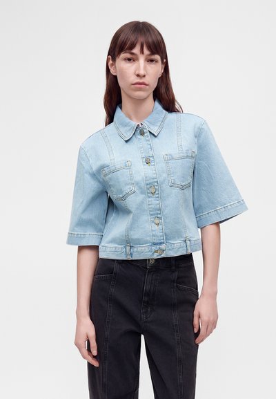 Jeune femme aux longs cheveux bruns portant une veste en jean bleu clair à manches courtes et un pantalon noir taille haute, debout devant un fond uni.