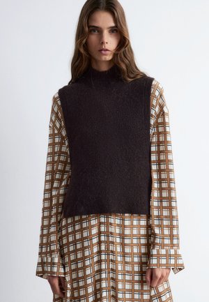 Gestricktes, dunkelbraunes ärmelloses Oberteil mit hohem Kragen über einem gemusterten, langärmeligen braunen, weißen und schwarzen karierten Kleid mit Knopfdetails.