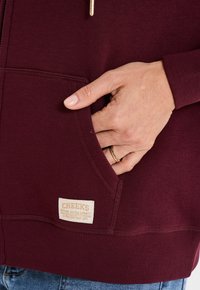Felpa con zip color borgogna, con polsini e orlo a costine, dotata di una tasca frontale e un'etichetta in tessuto con il logo "CREEKS" ricamato.