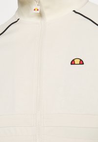 Veste zippée écrue avec bandes noires sur les épaules et petit logo en demi-cercle rouge et jaune sur la poitrine et sur la tirette.