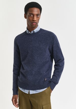 Giovane uomo che indossa un maglione girocollo blu navy sopra una camicia a righe blu con bottoni e pantaloni cargo oliva, che guarda dritto davanti a sé con un'espressione neutra.