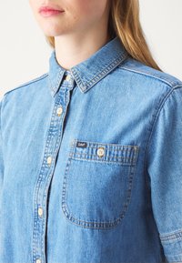 Femme portant une chemise en denim bleu clair avec fermeture boutonnée devant et une poche, affichant l'étiquette de la marque Lee sur la poche.