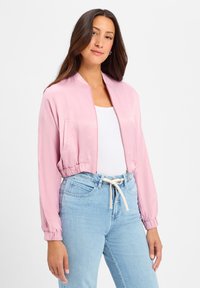 Lichtroze cropped bomberjack met elastische manchetten en taille, voorzien van een rits aan de voorkant en zijzakken, gedragen over een witte top en blauwe jeans.