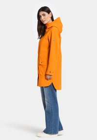 Ljus orange vattentät jacka med huva, knappränder och fickor, i kombination med ljusblå jeans och vita skor.