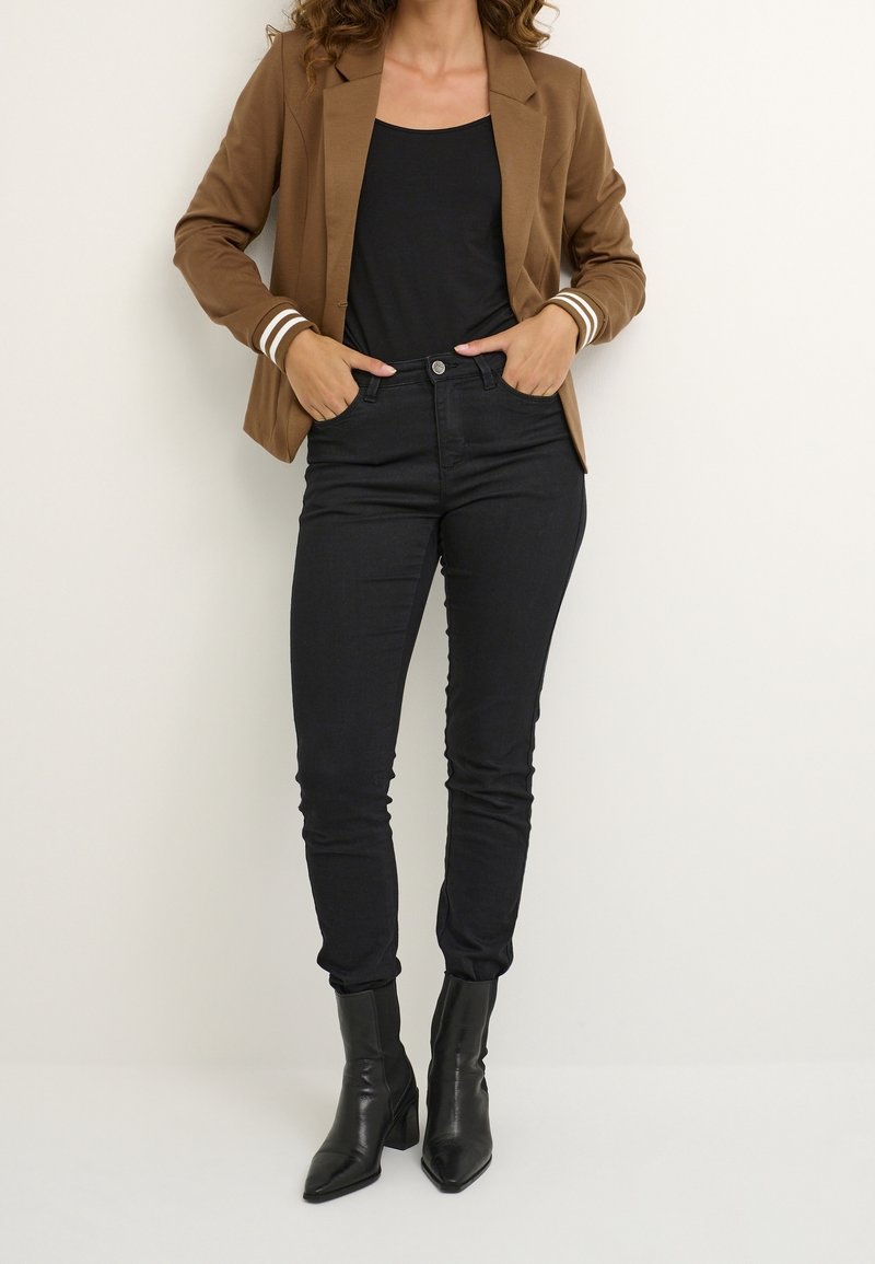 Femme portant un blazer marron avec des poignets rayés blancs, un top noir, un jean skinny noir et des bottines noires à bout pointu, debout les mains dans les poches.