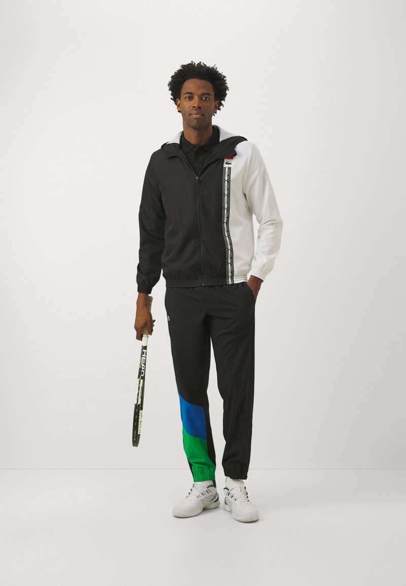 Uomo in giacca con zip bianca e nera e pantaloni neri con toppe blu e verdi, che tiene una racchetta da tennis su sfondo bianco.