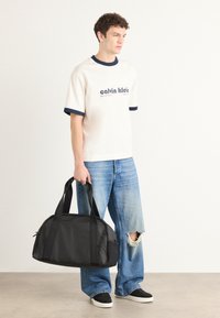 Svart duffelbag med doble håndtak på toppen, mykt materiale, rund form og glidelås. Modellen har på seg en kremfarget t-skjorte med marineblå detaljer og blå jeans.