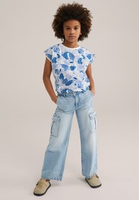 Camisa blanca de manga corta con un patrón floral azul, combinada con pantalones cargo azul claro que tienen bolsillos laterales y un corte relajado. Calzado marrón.