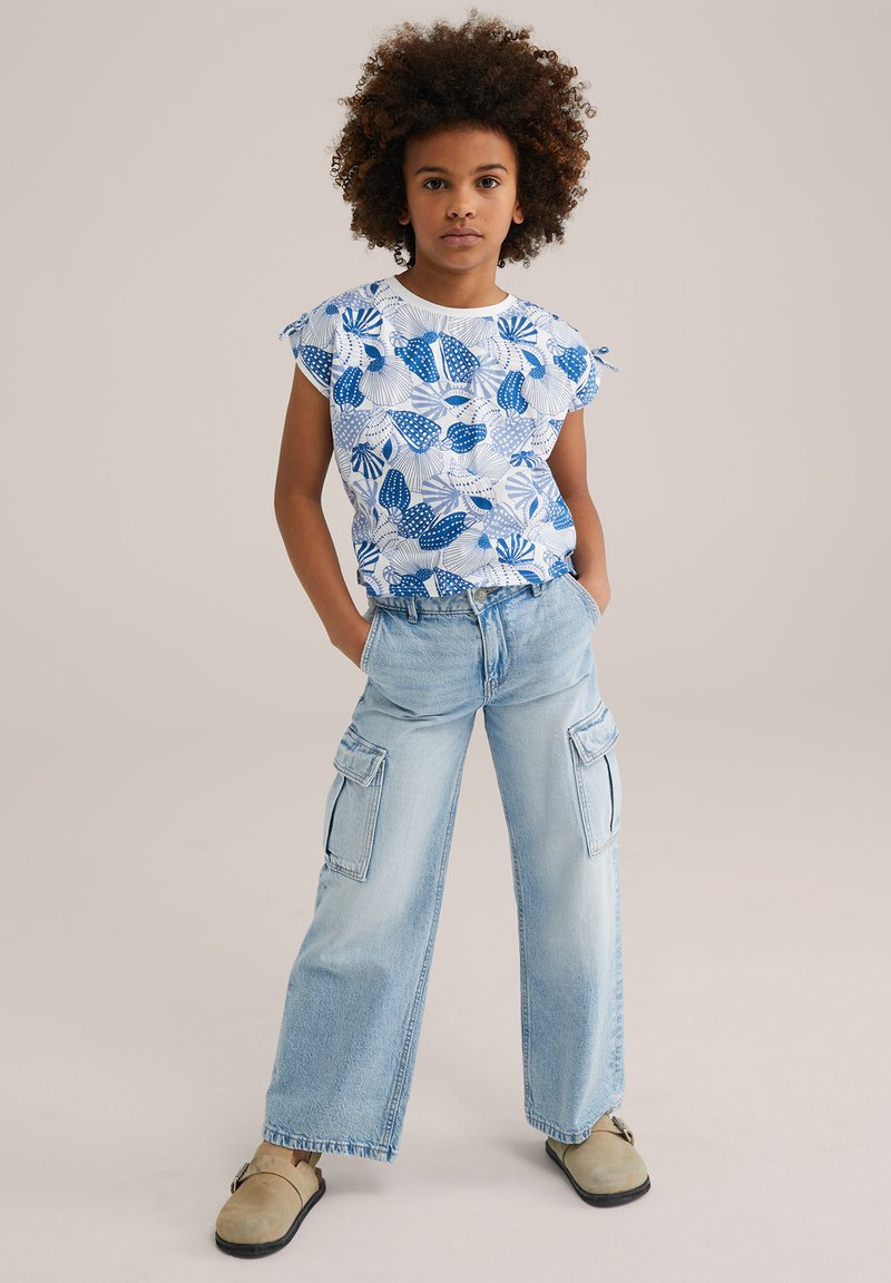 Camisa blanca de manga corta con un patrón floral azul, combinada con pantalones cargo azul claro que tienen bolsillos laterales y un corte relajado. Calzado marrón.