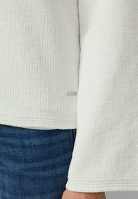 Biały sweter o fakturze z prążkowanej tkaniny, z widoczną metką marki przy dolnym brzegu. Charakteryzuje się szerokimi rękawami i luźnym krojem, noszony zestawiony z dżinsem.