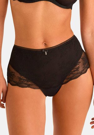 Femme portant des sous-vêtements noirs en dentelle taille haute et un soutien-gorge assorti, montrant le ventre et le haut des cuisses sur un fond uni.