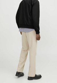 Svart bomberjacka med elastiska ärmslut, lager på lager över en randig skjorta. Beiga byxor och svarta skor fulländar outfiten.