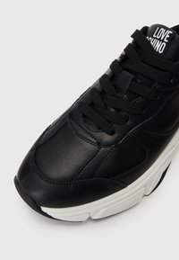 Melnas ādas sporta kedas ar baltu zoli, melniem šņorēm un uzrakstu "LOVE MOSCHINO" uz mēles, attēlots no augšpuses priekšējā leņķī.