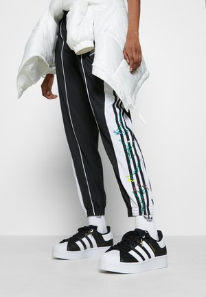 Zwart-witte trainingsbroek met bloemmotief, witte pufferjas over de schouder gedrapeerd, en zwarte Adidas sneakers met witte accenten en een shell-toe.