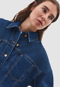 Ung kvinna med långt brunt hår klädd i en blå jeansjacka med metallknappar och gyllene hoops, som tittar åt sidan.