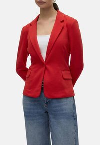 Blazer structuré rouge avec revers, fermeture à un bouton unique et poches avant. Porté par-dessus un haut blanc et un jean large bleu.