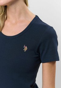 Krátky rukáv, námornícky modrý rebrovaný tričko s okrúhlym výstrihom a vyšívaným logom na hrudi. Hladká textúra so priliehavým strihom.