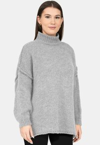 Vrouw met een losse, lichtgrijze gebreide coltrui en zwarte broek, staand tegen een effen lichte achtergrond.