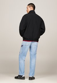 Zwarte bomberjack met hoge kraag, voorzien van elastische manchet en zoom. G gedragen met lichtblauwe jeans en zwarte sneakers.