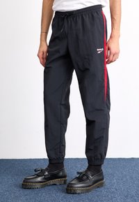 Čierne športové nohavice s červenými bočnými pruhmi, elastickým pásom a manžetami, vyrobené z ľahkej tkaniny. Obsahujú logo Reebok na stehne.