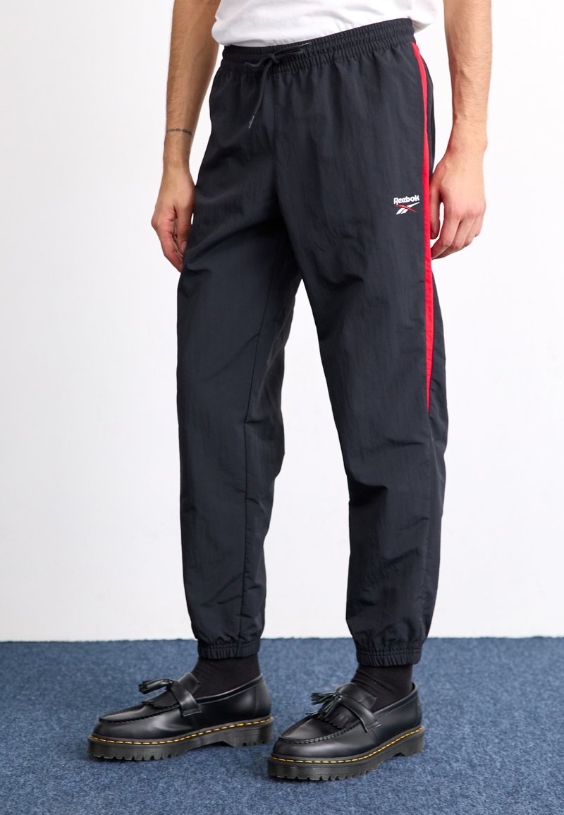 Čierne športové nohavice s červenými bočnými pruhmi, elastickým pásom a manžetami, vyrobené z ľahkej tkaniny. Obsahujú logo Reebok na stehne.
