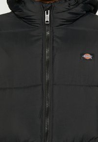 Black puffer jacket featuring a zip-up front, high collar, and horizontal quilted pattern. Includes a small logo patch on the chest.