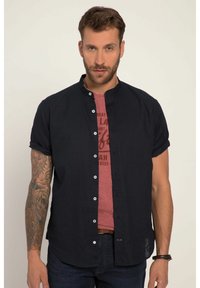 Camicia blu navy a maniche corte con colletto a banda, con bottoni bianchi, indossata sopra una maglietta rossa con grafica, abbinata a jeans scuri.