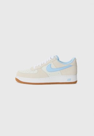 Tenisky Nike Air Force 1 majú béžovú semišovú vrchnú časť, svetlomodré detaily, biele šnúrky a gumovú podrážku s „AIR“ brandingom.