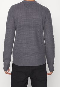 Osoba w szarej, długosleeve'owej, strukturalnej bluzie i czarnych spodniach, widoczna z tyłu na jednolitej białej stronie.