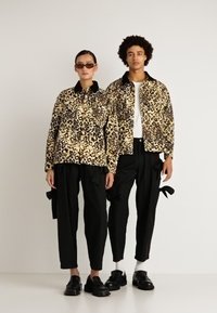 Két modell, akik egymáshoz illő leopárdmintás dzsekiket viselnek, fekete cargo nadrággal és kötőkkel, fekete cipővel; az egyik napszemüveget visel, sima háttér előtt állnak.