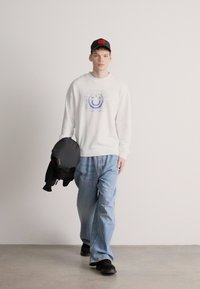 Λευκό φούτερ με μπλε γραφικό χαμογελαστό πρόσωπο, ανοιχτόχρωμα μπλε τζιν, και μαύρα παπούτσια χωρίς κορδόνια, κρατώντας μαύρο μπουφάν, φοράει μαύρο καπέλο.