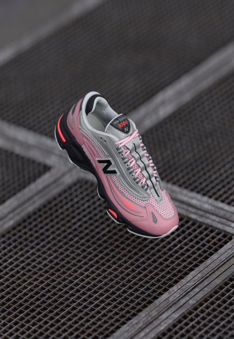 Rozi i sivi New Balance 1000 tenisica s crnom podlogom i rozim vezicama lebdi iznad tamnog metalnog rešetkastog poda.