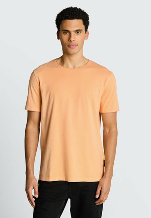 Homme aux cheveux courts et bouclés, portant un t-shirt uni pêche à manches courtes et un pantalon noir, face à un fond gris clair.