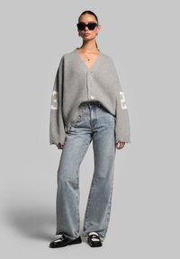 Gray ribbed cardigan with a V-neck, buttons, and white number accents. Paired with high-waisted blue jeans and black loafers.