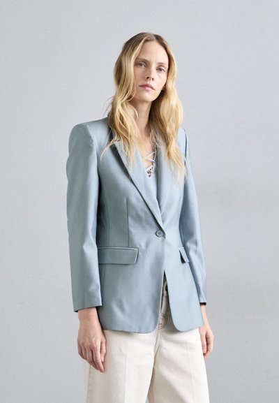 Donna con lunghi capelli biondi che indossa un blazer su misura blu chiaro e pantaloni a gamba larga color crema, in un contesto grigio neutro.