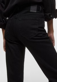 Jeans in denim nero con una texture liscia, caratterizzati da un design a cinque tasche, dettagli cuciti e una patch in pelle nera con logo sul retro.
