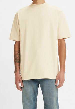 Homme portant une chemise beige unie à manches courtes avec un col montant et un jean bleu, debout devant un fond blanc.
