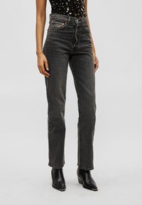 Γυναίκα με ψηλόμεσα ξεθωριασμένα μαύρα τζιν και μαύρα μποτάκια με μυτερή μύτη, σε απλό φωτεινό φόντο.