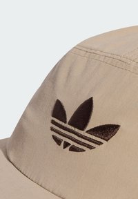 Gorra de tela beige con un logo bordado en marrón oscuro que presenta tres hojas y rayas horizontales, destacando una textura suave y una forma curvada.