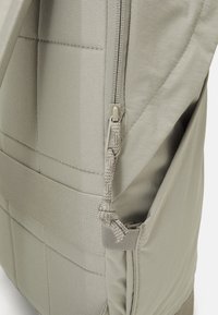 Gros plan d'une poche latérale beige d'un sac à dos avec un curseur de fermeture éclair texturé et des panneaux en tissu cousus.