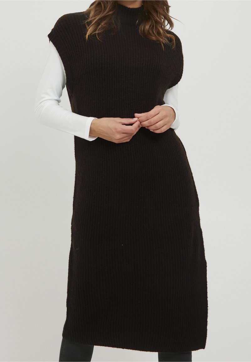 Femme portant une robe noire côtelée sans manches par-dessus un haut blanc à manches longues, mains jointes à la taille, debout devant un fond uni.