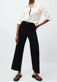 Los vaqueros negros de pierna ancha presentan un dobladillo deshilachado, combinados con una camisa de botones en color crema. El conjunto se complementa con sencillas sandalias negras.