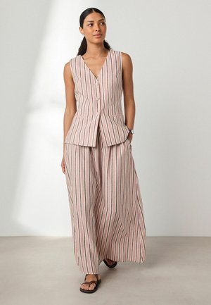 Frau stehend, trägt blassrosa ärmellose gestreifte Weste und weit geschnittene passende Hose mit schwarzen Sandalen, vor schlichter hellem Hintergrund.