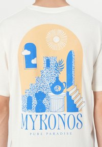 Witte katoenen T-shirt met een blauw en oranje grafisch ontwerp met trappen, vazen, wolken en de tekst "MYKONOS PURE PARADISE."