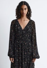 Schwarzes Blumenkleid mit tiefem V-Ausschnitt, langen transparenten Ärmeln, gerafftem Taillenbereich und fließender Silhouette, die mit kleinen bunten Blumenmustern verziert ist.