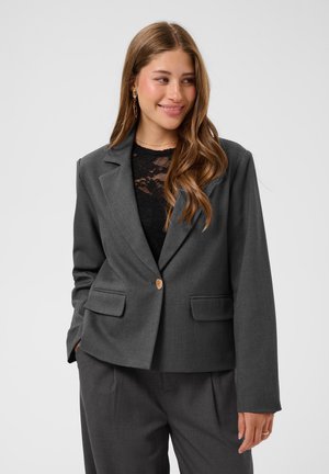 Jeune femme aux longs cheveux portant une veste de costume gris foncé sur un top en dentelle noire, souriante et regardant sur le côté.