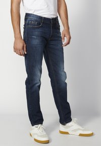 Sötétkék farmernadrág, slim fit fazonban, öt zsebbe és kontrasztos varrással. Fehér sportcipővel viselve.